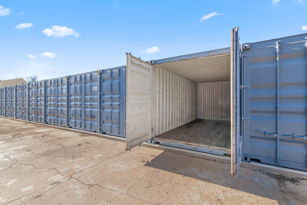 Open shipping container with empty interior, flanked by closed containers, under clear blue sky on a concrete surface.