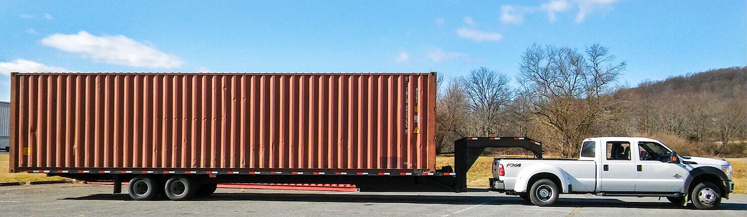 intro-bg1 A white pickup truck is hauling a long, brown shipping container on a trailer in a paved area with trees and hills in the background.