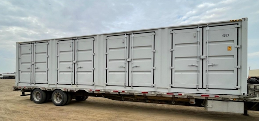 Picture of New Open Side 40′ Unit Shipping Container A large white trailer with multiple closed cargo doors is parked on a dirt lot under a cloudy sky.