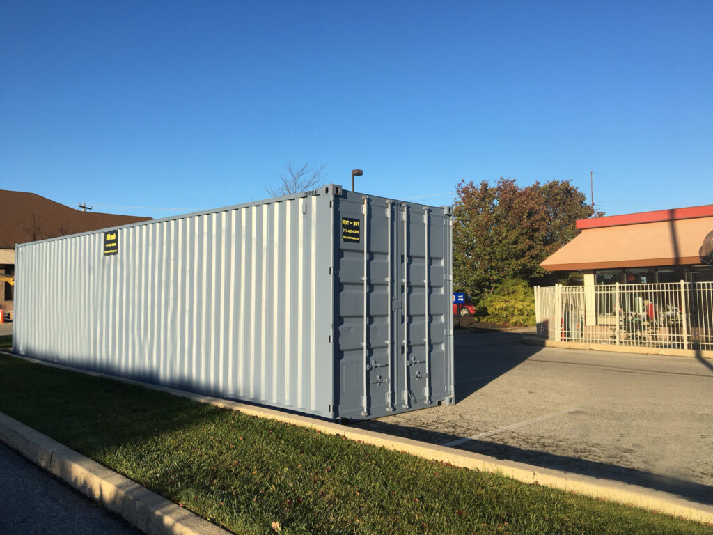 Portable Storage Delivery – no shadow A large gray shipping container sits on pavement near a grassy area, with buildings and a fence visible in the background under a clear blue sky.