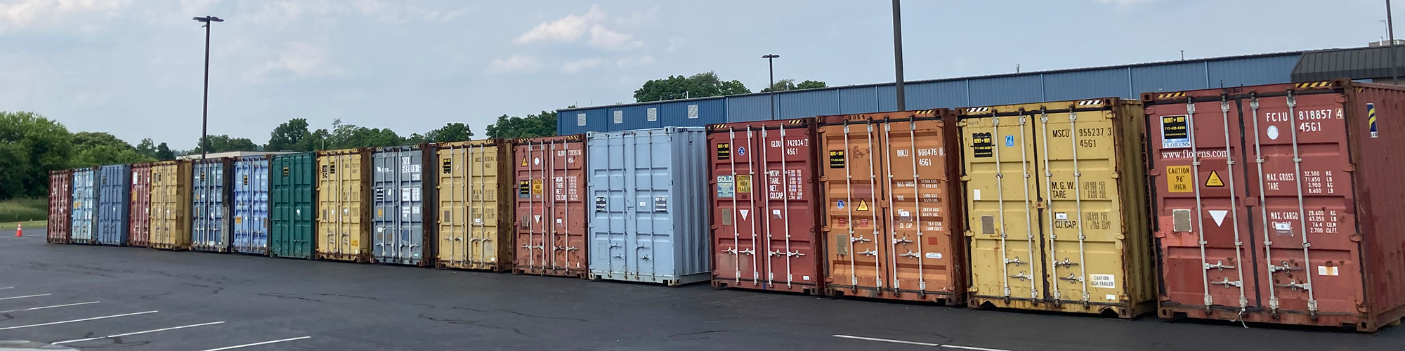 A row of colorful shipping containers lined up in a parking lot near a building and trees under a cloudy sky.