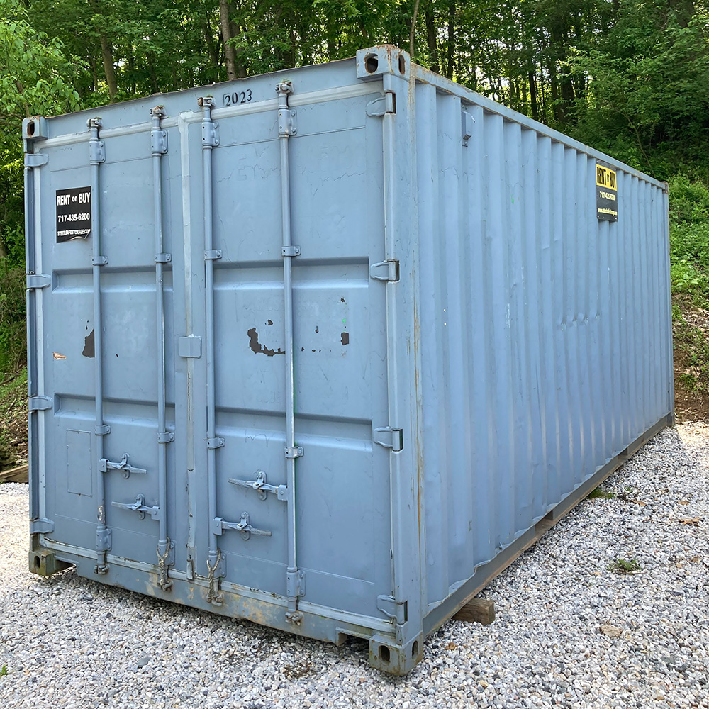 20ft Portable Container A large blue metal shipping container sits on a gravel surface, surrounded by trees and grass, with signs for rental and purchase on its doors.