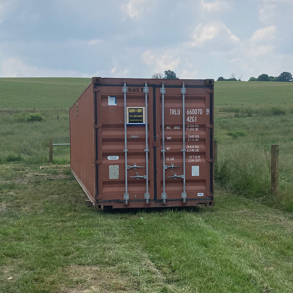 Agriculture Residential Storage Container A brown shipping container sits on grass in a rural area, with a field and cloudy sky in the background.