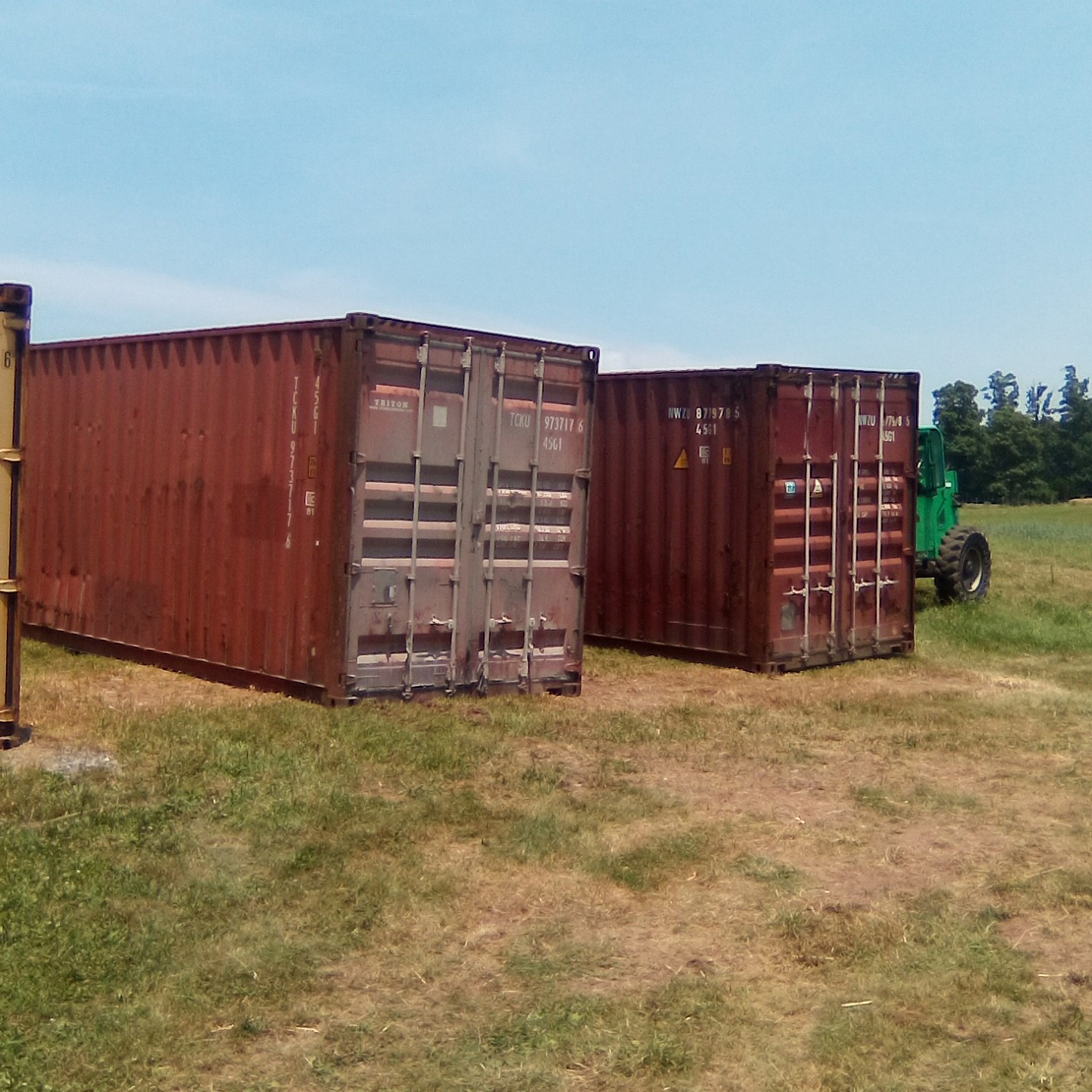 Agriculture Storage Container Two large red shipping containers are placed on a grassy field with a green tractor partially visible behind them.