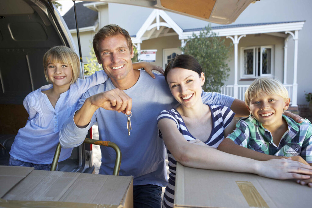 A smiling family of four stands by a moving van with cardboard boxes; the father holds house keys in front of a suburban home.