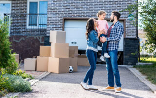 A family stands together in front of a house with stacked moving boxes on the driveway, suggesting they are moving in or out.