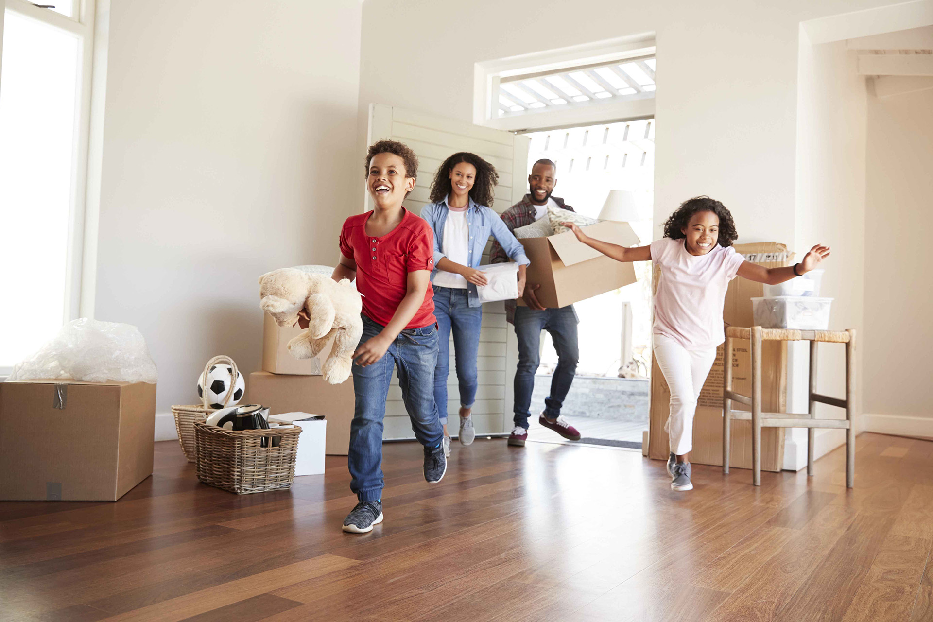 A family of four enters a new home carrying boxes and belongings, with two children running ahead and parents smiling in the background.