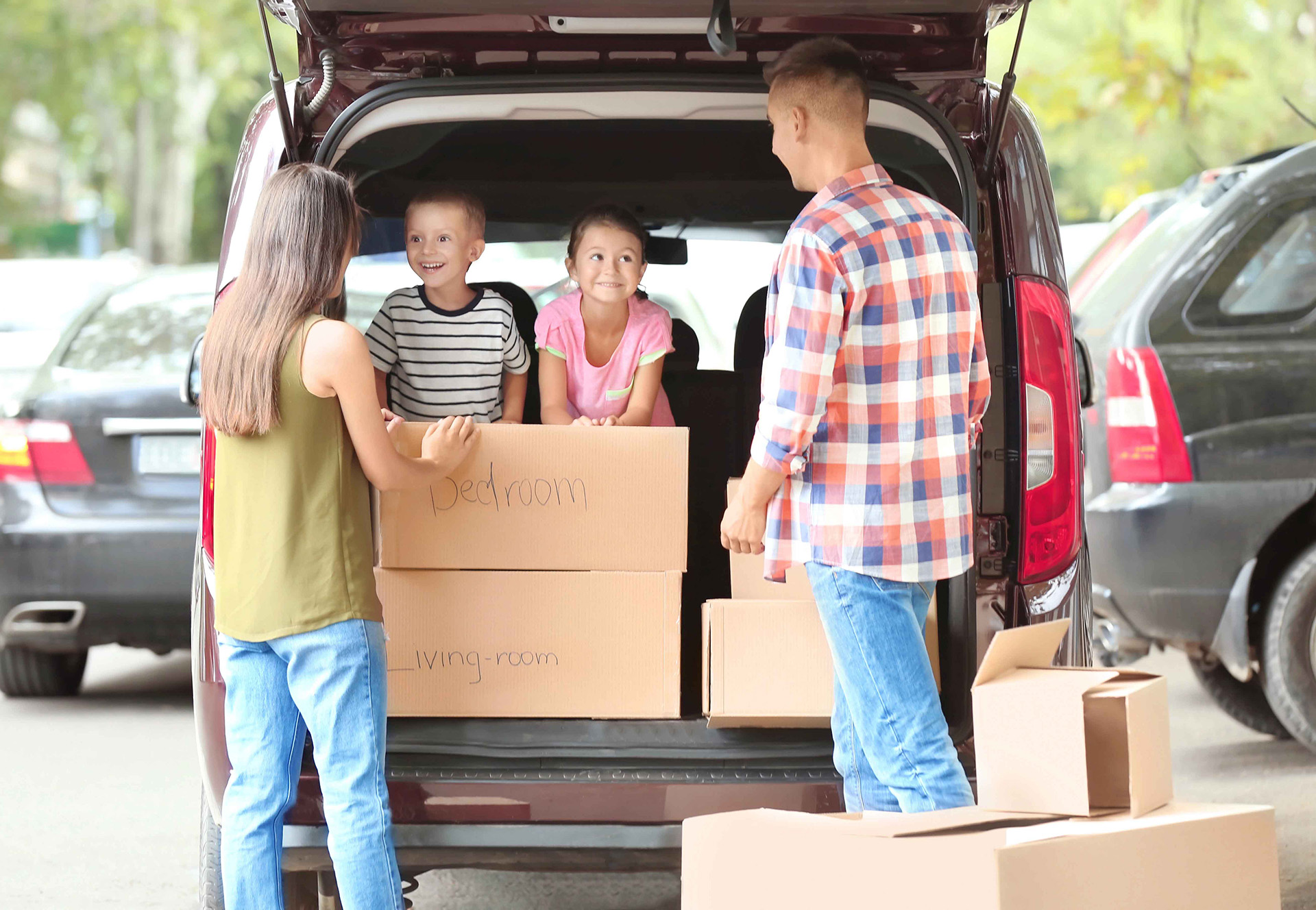 Two adults and two children load cardboard boxes labeled "bedroom" and "living room" into the back of a minivan in a parking lot.