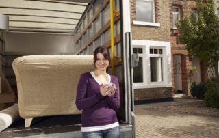A woman holding a mug stands by a moving truck loaded with furniture outside a brick house with the number 62 on the front.
