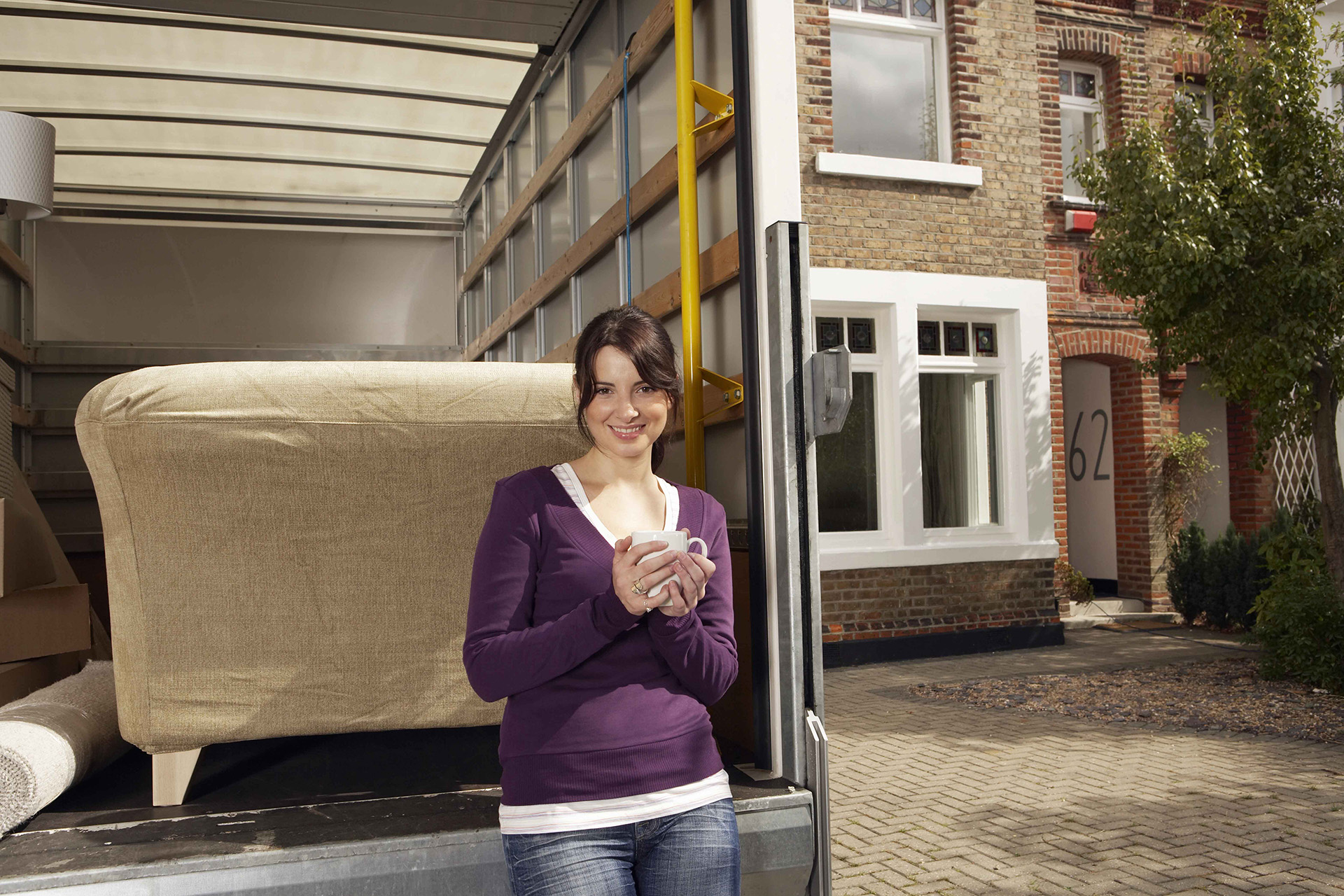 A woman holding a mug stands by a moving truck loaded with furniture outside a brick house with the number 62 on the front.