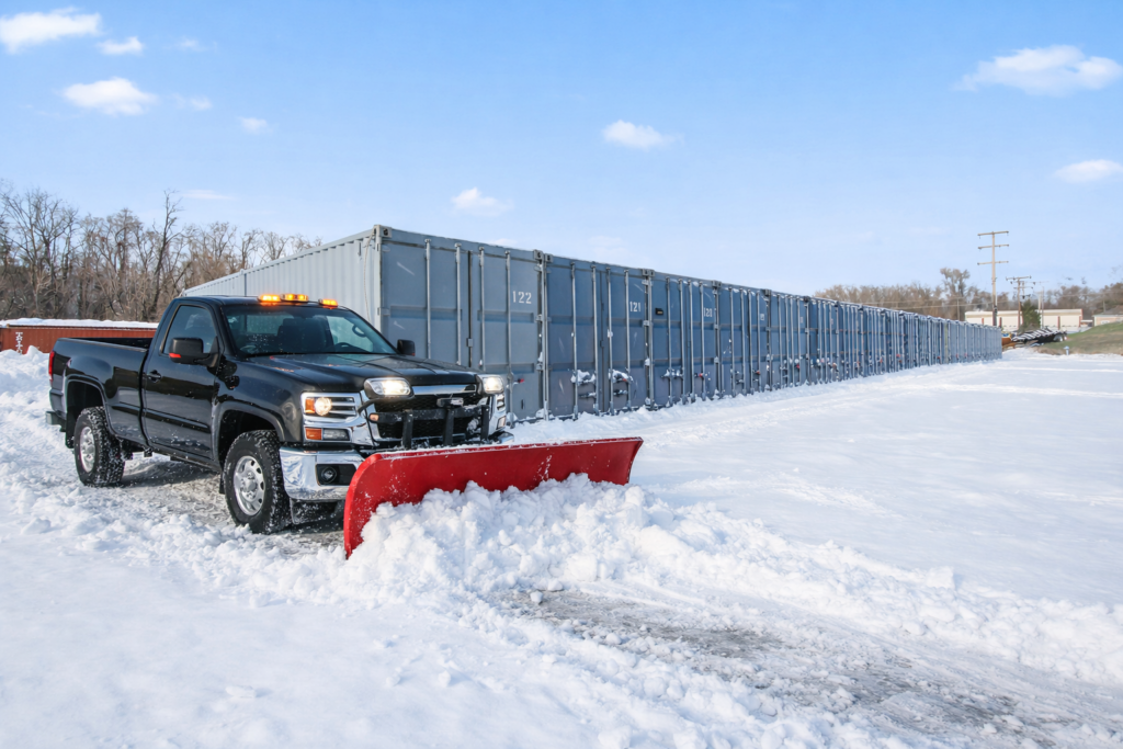 A black pickup truck with a snowplow clears snow in front of a row of large gray shipping containers on a bright winter day.
