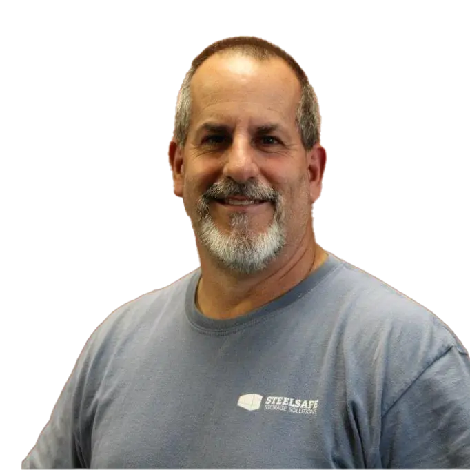 A man with short hair and a gray beard is wearing a light blue “Steelsafe Storage Solutions” T-shirt, smiling against a plain white background.