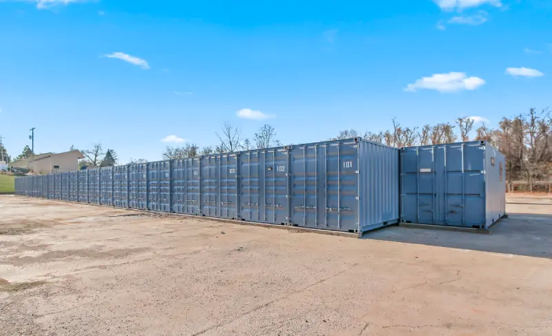 A row of large blue shipping containers sits on a paved lot under a clear sky, with trees and a building in the background.